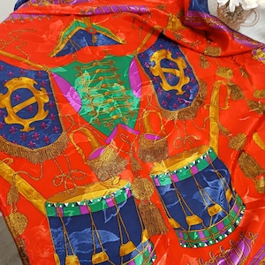 May include: A vibrant red silk scarf with a repeating pattern of drums, flags, and decorative elements in gold, blue, green, and purple. The scarf has a glossy sheen and a detailed design, suitable for fashion or decorative use.