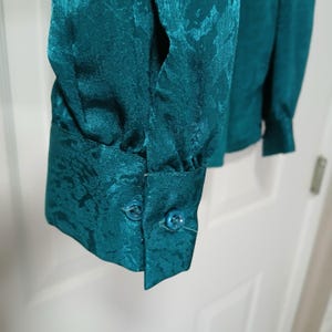 Peut inclure: Gros plan sur une manche de chemisier de couleur sarcelle. Le tissu pr&eacute;sente un motif subtil en relief. Le poignet comporte deux petits boutons ronds. Le chemisier semble &ecirc;tre fait d'une mati&egrave;re soyeuse.