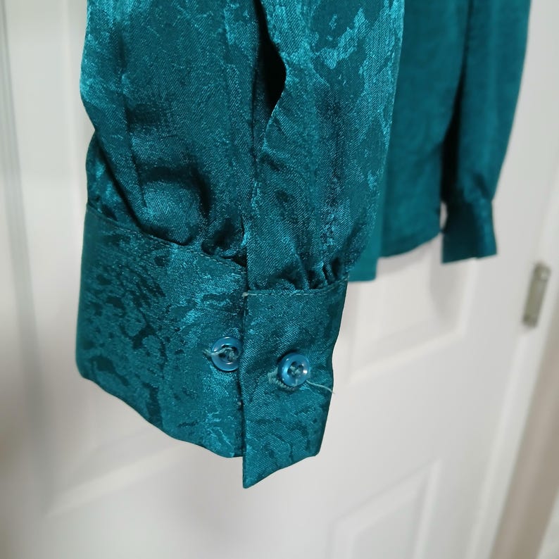 Peut inclure: Gros plan sur une manche de chemisier de couleur sarcelle. Le tissu pr&eacute;sente un motif subtil en relief. Le poignet comporte deux petits boutons ronds. Le chemisier semble &ecirc;tre fait d'une mati&egrave;re soyeuse.