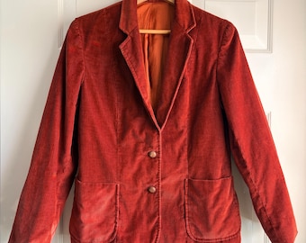 May include: A burnt orange corduroy blazer featuring a notched lapel, two front patch pockets, and a two-button closure. The jacket is fully lined with a matching orange fabric and is displayed on a wooden hanger against a white door.