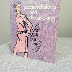 May include: A hardcover book titled Pattern Drafting and Dressmaking by Dorothy Moore. The cover features a purple and pink textured background with black line-art illustrations of two figures wearing clothing, one standing and one sitting.