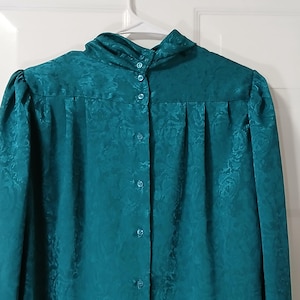 Peut inclure: Chemisier boutonn&eacute; de couleur teal avec un col montant. Le chemisier pr&eacute;sente un motif floral subtil et est suspendu &agrave; un cintre blanc contre une porte blanche. Le chemisier &agrave; manches longues a des boutons sur le devant et des plis aux &eacute;paules.