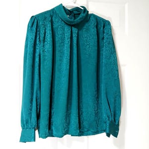 Peut inclure: Chemisier de couleur teal avec un col montant fronc&eacute; et des manches longues. Le tissu pr&eacute;sente un motif floral subtil. Les poignets sont boutonn&eacute;s. Le chemisier est suspendu &agrave; un cintre blanc contre une porte blanche.