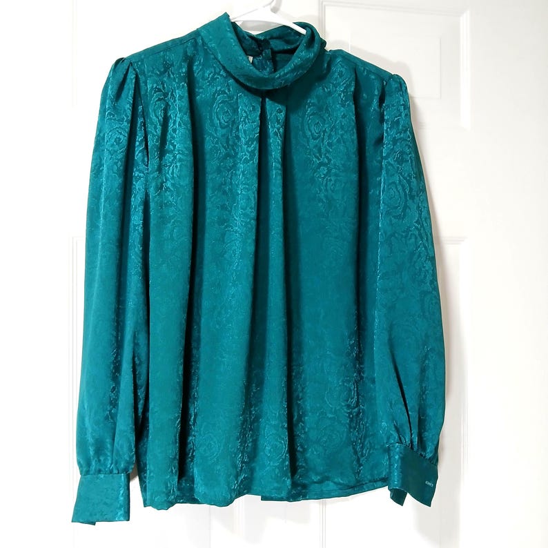 Peut inclure: Chemisier de couleur teal avec un col montant fronc&eacute; et des manches longues. Le tissu pr&eacute;sente un motif floral subtil. Les poignets sont boutonn&eacute;s. Le chemisier est suspendu &agrave; un cintre blanc contre une porte blanche.