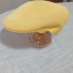 Puede incluir: Gorra de estilo newsboy de punto amarillo claro. La gorra se exhibe sobre un pedestal de vidrio transparente y decorativo. Tiene una corona redondeada y una visera corta y r&iacute;gida. La tela de punto tiene una textura.