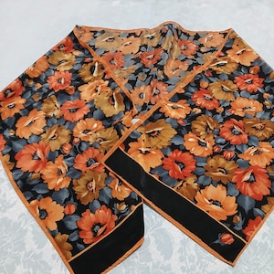 May include: A floral scarf featuring a pattern of orange and yellow flowers on a black background. The scarf has a black border with an orange trim. The scarf is made of a silky material and is folded in a decorative manner.
