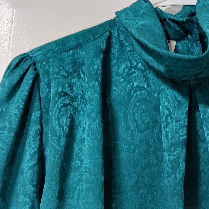 Peut inclure: Chemisier de couleur teal avec un col montant et des manches bouffantes. Le tissu pr&eacute;sente un motif floral subtil. Le chemisier est fait d'une mati&egrave;re brillante, et le gros plan met en valeur la texture et le design du v&ecirc;tement.