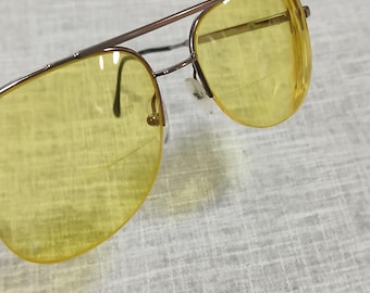 Puede incluir: Primer plano de gafas de sol de aviador con lentes de color amarillo y montura de metal marrón. Las lentes son de color amarillo brillante y la montura tiene un diseño de doble puente. Las almohadillas nasales son blancas.