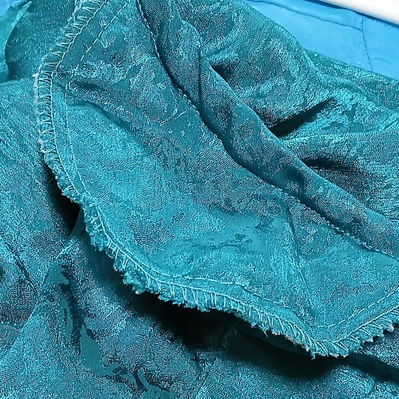Peut inclure: Gros plan sur un tissu turquoise &agrave; motif floral. Le tissu est pli&eacute;, r&eacute;v&eacute;lant une surface textur&eacute;e et un bord cousu. La couleur est un turquoise vif, avec de subtiles variations de tons.