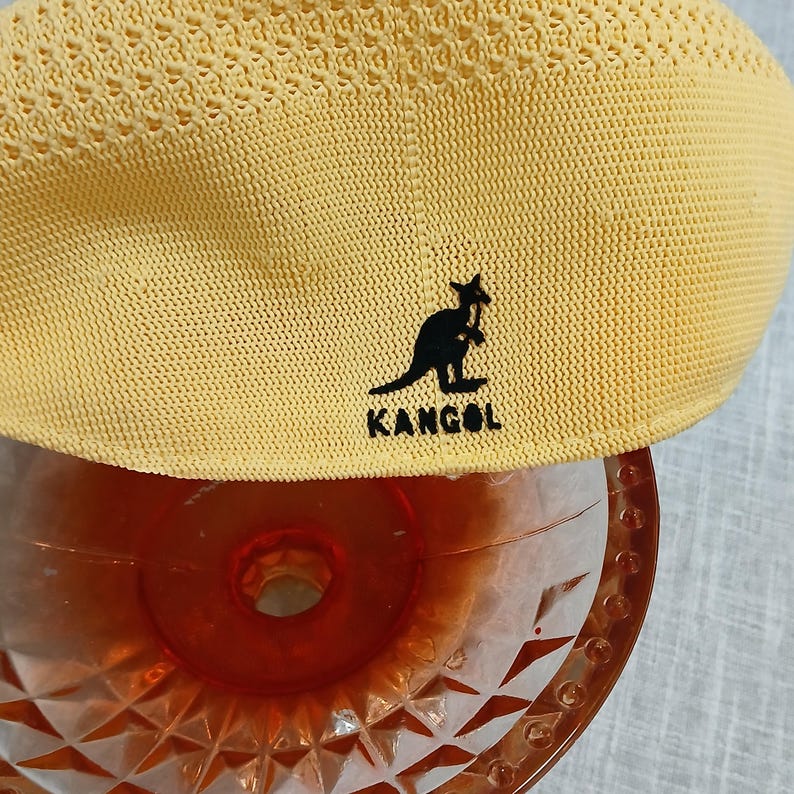 Puede incluir: Una boina Kangol amarilla con un dise&ntilde;o de punto texturizado. La boina presenta el logotipo negro de Kangol. El sombrero est&aacute; sobre un vaso decorativo.