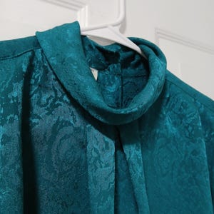 Peut inclure: Chemisier de couleur teal avec un col roul&eacute; haut. Le tissu pr&eacute;sente un motif floral discret. Le chemisier est suspendu &agrave; un cintre blanc sur fond blanc.