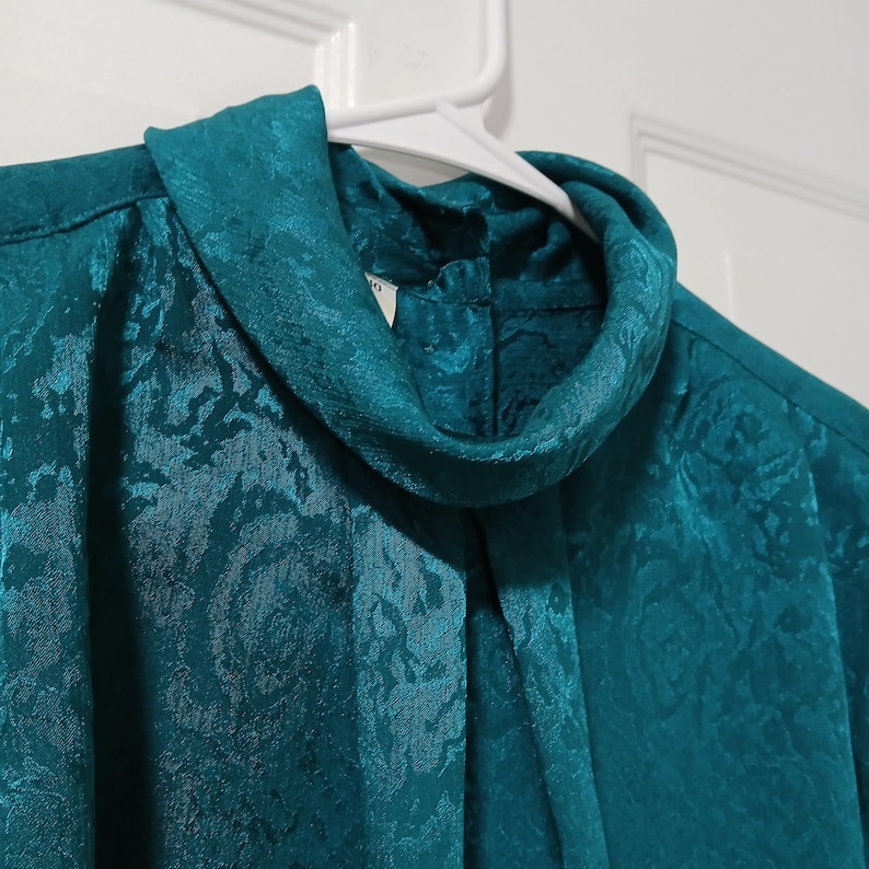 Peut inclure: Chemisier de couleur teal avec un col roul&eacute; haut. Le tissu pr&eacute;sente un motif floral discret. Le chemisier est suspendu &agrave; un cintre blanc sur fond blanc.