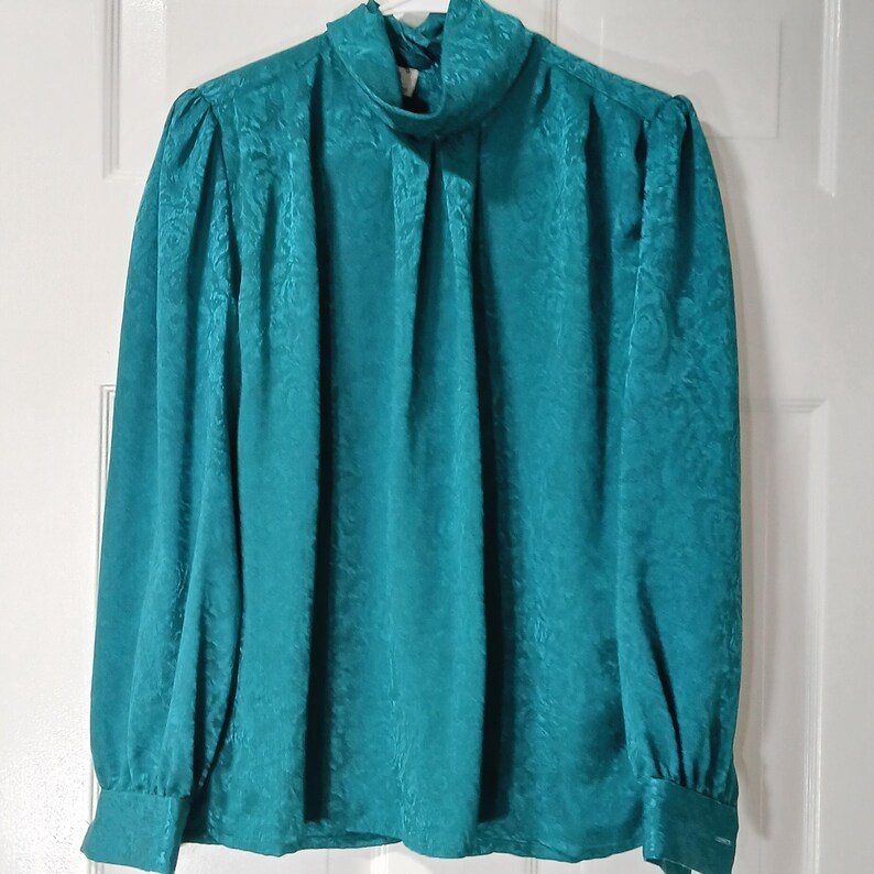 Peut inclure: Chemisier de couleur teal avec un col montant fronc&eacute; et des manches longues. Le tissu pr&eacute;sente un motif floral subtil en relief. Les manches ont un l&eacute;ger effet bouffant aux &eacute;paules et des poignets boutonn&eacute;s. Le chemisier est suspendu contre une porte blanche.