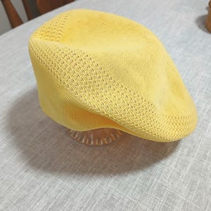 Puede incluir: Gorra plana de punto amarillo brillante con una corona redondeada y una visera corta y r&iacute;gida. La gorra tiene un dise&ntilde;o texturizado de tejido abierto y est&aacute; sobre un soporte decorativo transparente. Un accesorio elegante.