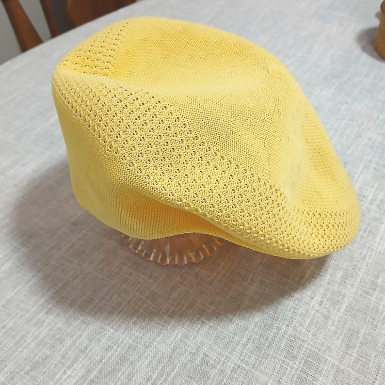 Puede incluir: Gorra plana de punto amarillo brillante con una corona redondeada y una visera corta y r&iacute;gida. La gorra tiene un dise&ntilde;o texturizado de tejido abierto y est&aacute; sobre un soporte decorativo transparente. Un accesorio elegante.