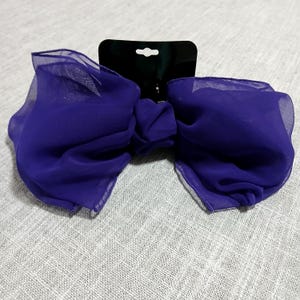 May include: A large, vibrant purple bow hair accessory. The bow is made of a sheer, flowing fabric, and is attached to a black tag. The bow is a deep shade of purple, and the fabric has a slight sheen.