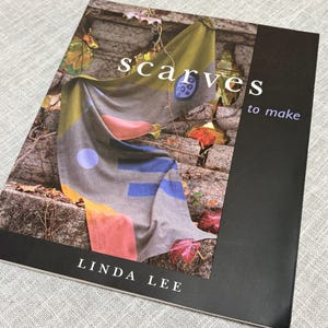 May include: A softcover book titled Scarves to Make by Linda Lee. The cover features a photograph of a grey scarf with yellow, pink, and blue geometric shapes draped over a stone wall with autumn leaves. The text is in white and blue.