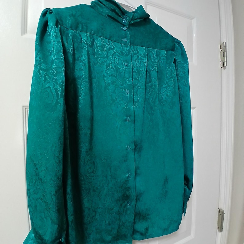 Peut inclure: Un chemisier boutonn&eacute; de couleur sarcelle avec des manches longues et un col montant. Le tissu pr&eacute;sente un motif floral subtil. Le chemisier est suspendu &agrave; une porte blanche.