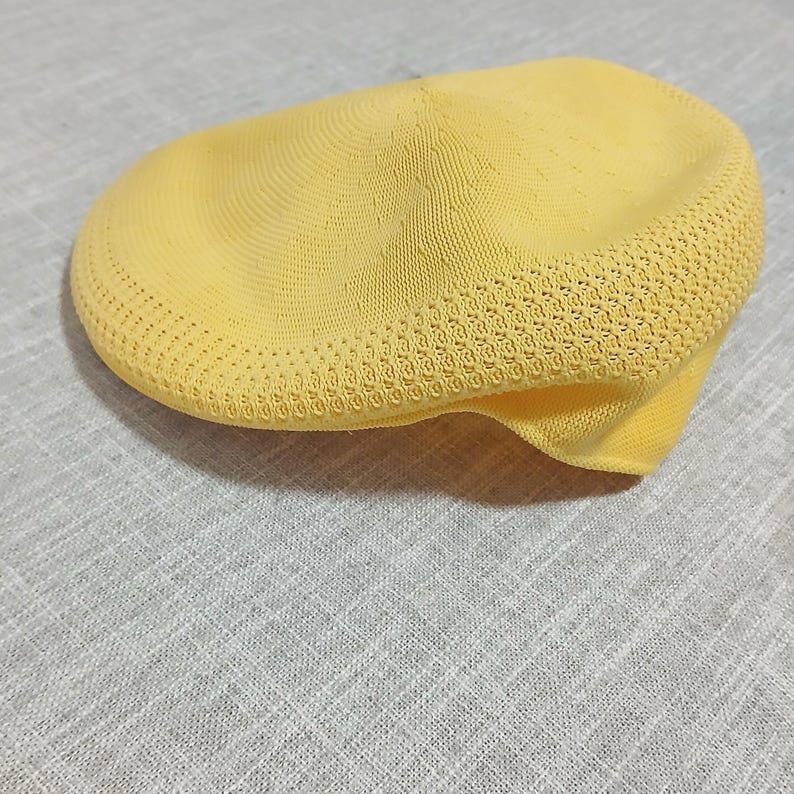 Puede incluir: Gorra plana amarilla, tejida con un patr&oacute;n texturizado alrededor del borde y una corona lisa. El sombrero tiene una visera corta y r&iacute;gida. Este sombrero ligero est&aacute; probablemente dise&ntilde;ado para uso informal y es un estilo cl&aacute;sico.