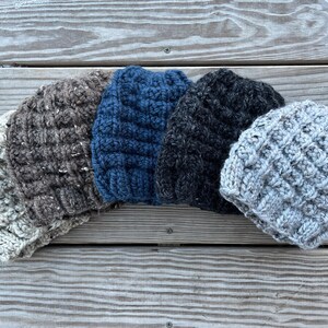 May include: Four knitted hats in different colors and textures. The hats are arranged in a semi-circle on a wooden surface. The hats are made of a chunky yarn and have a cable knit pattern.