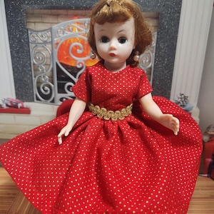 May include: A vintage doll wearing a red dress with gold polka dots and a gold belt. The doll has short brown hair, pearl earrings, and is posed in front of a fireplace.