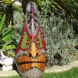 Op de afbeelding: Een houten tiki-masker dat rood, groen en bruin is geschilderd. Het masker heeft een brede, tanden glimlach en is versierd met geometrische patronen.