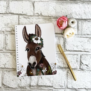 May include: A white spiral bound notebook featuring an illustration of a brown donkey wearing a white flower crown and surrounded by colourful flowers.