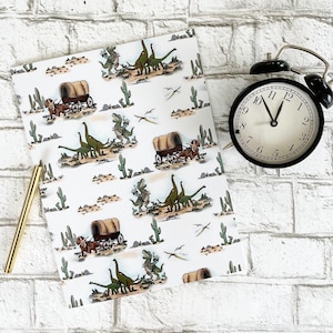May include: A white paper with a repeating pattern of dinosaurs, covered wagons, and cacti. A black alarm clock with a white face and gold pen are next to the paper. The clock shows the time is approximately 11:00.