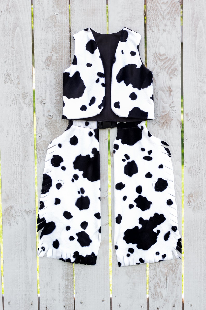 May include: A black and white cow print vest and chaps costume. The vest has a black lining and the chaps have fringe along the bottom.