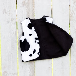 May include: A black and white cow print vest with a black fabric lining. The vest is hanging on a white wooden fence.