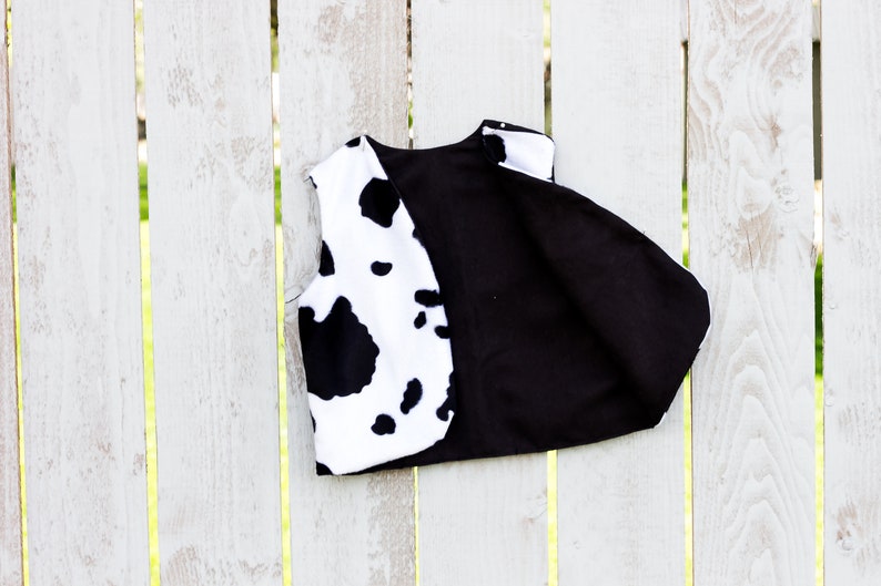 May include: A black and white cow print vest with a black fabric lining. The vest is hanging on a white wooden fence.