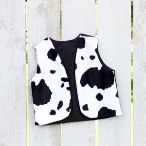 May include: A black and white cow print vest with a black lining. The vest is hanging on a white wooden fence.