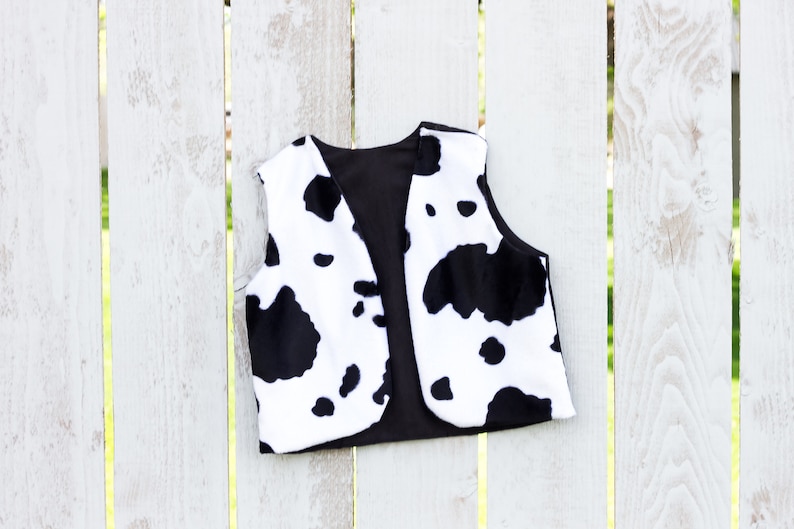 May include: A black and white cow print vest with a black lining. The vest is hanging on a white wooden fence.