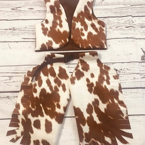 May include: A brown and white cow print vest and chaps with fringe. The vest has a brown lining and a buckle on the back. The chaps have a brown belt with a buckle.