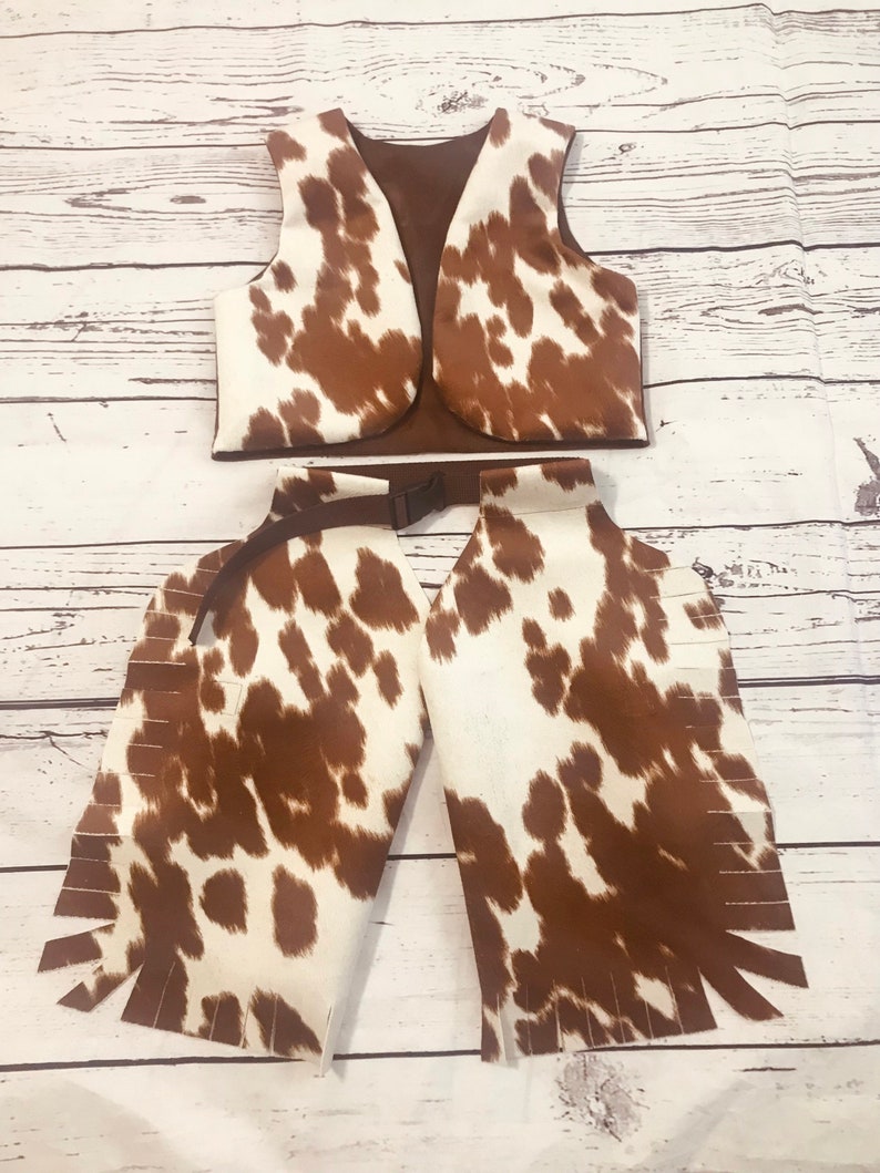 May include: A brown and white cow print vest and chaps with fringe. The vest has a brown lining and a buckle on the back. The chaps have a brown belt with a buckle.