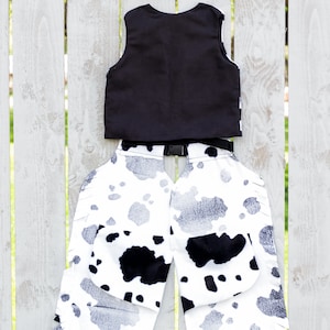 May include: Black vest and white and black cow print chaps with fringe. The chaps have a black belt with a buckle.