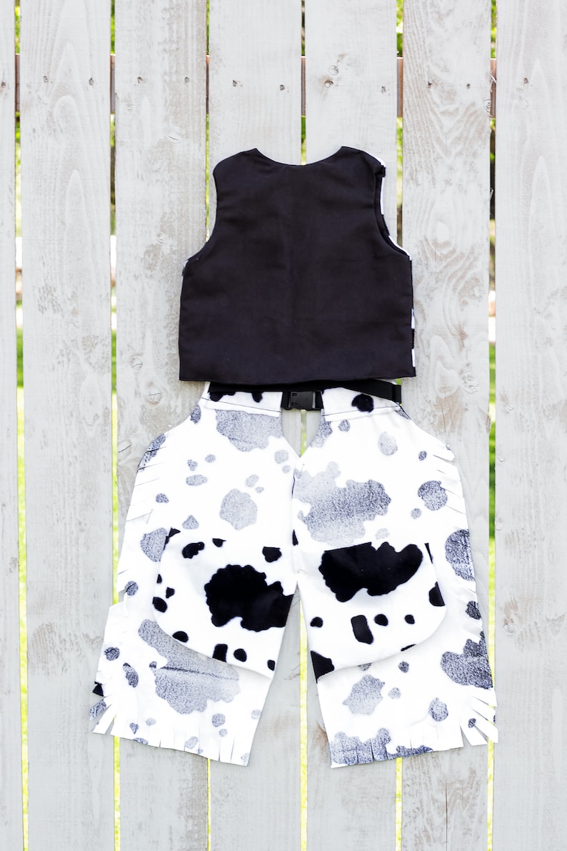 May include: Black vest and white and black cow print chaps with fringe. The chaps have a black belt with a buckle.
