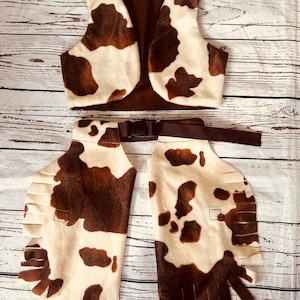 Könnte beinhalten: Cowboy-Kostüm-Set mit Weste, Gürtel und Chaps. Die Weste und Chaps sind cremefarben mit braunen Kuhfellflecken. Der Gürtel ist braun. Die Chaps haben einen Fransenrand. Das Kostüm liegt auf einer Holzoberfläche.
