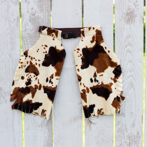 May include: Cow print chaps with brown fringe and a brown adjustable buckle.
