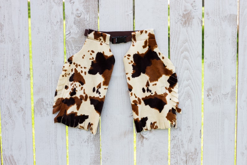 May include: Cow print chaps with brown fringe and a brown adjustable buckle.