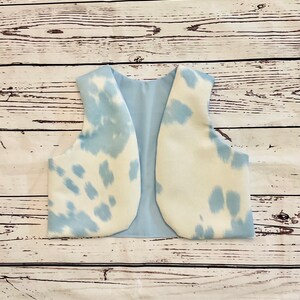 May include: A light blue and white cow print vest. The vest has a light blue interior lining. The exterior features a white base with irregular light blue spots. The vest is open in the front and has a rounded bottom.