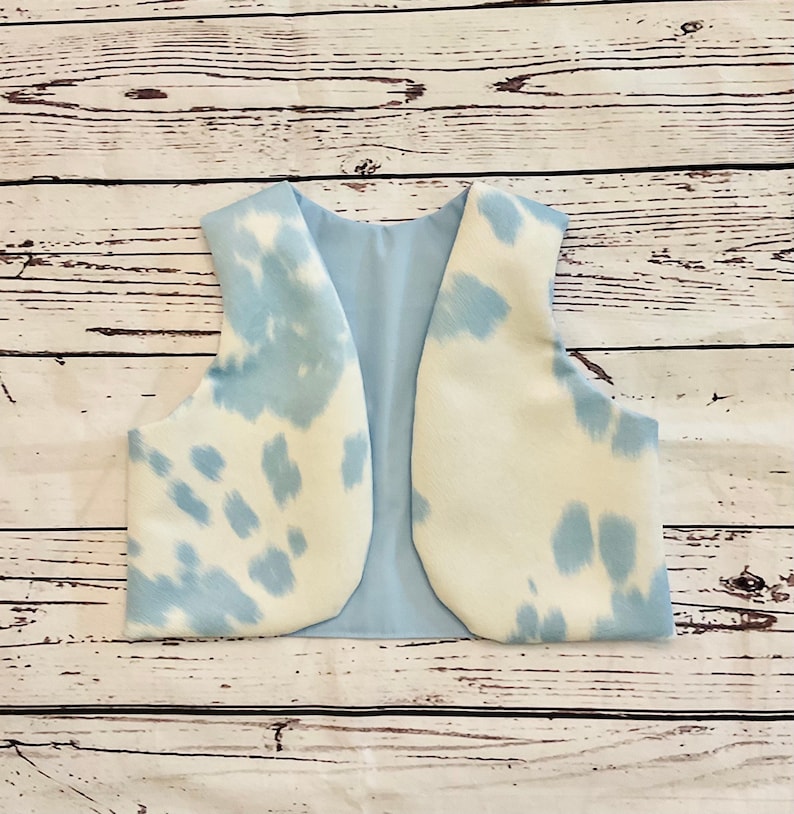 May include: A light blue and white cow print vest. The vest has a light blue interior lining. The exterior features a white base with irregular light blue spots. The vest is open in the front and has a rounded bottom.