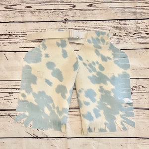May include: Light blue and white cow print chaps with fringe detailing. The chaps have a white belt with a buckle. The chaps are made of a soft material and are designed for children.
