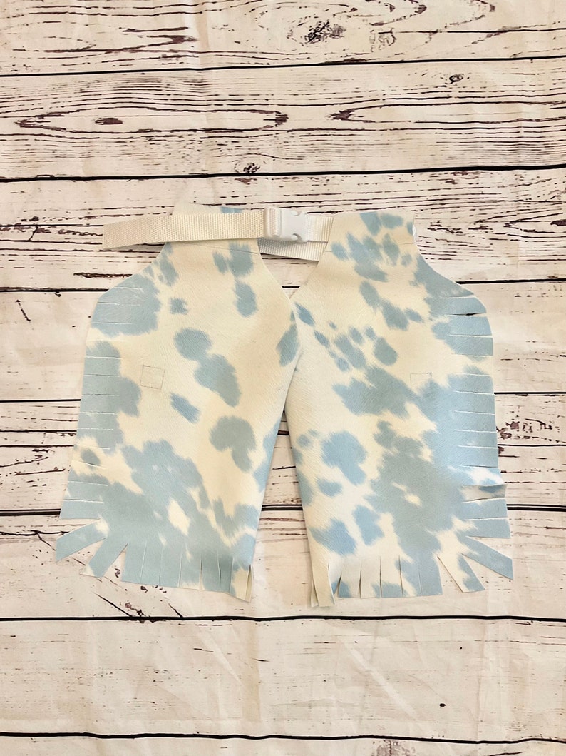 May include: Light blue and white cow print chaps with fringe detailing. The chaps have a white belt with a buckle. The chaps are made of a soft material and are designed for children.