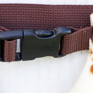 May include: A brown adjustable strap with a black plastic buckle. The strap is made of woven fabric.