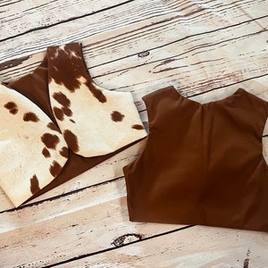 May include: Two brown vests, one with a white and brown cowhide pattern. The vests are laid flat on a light brown wooden surface.