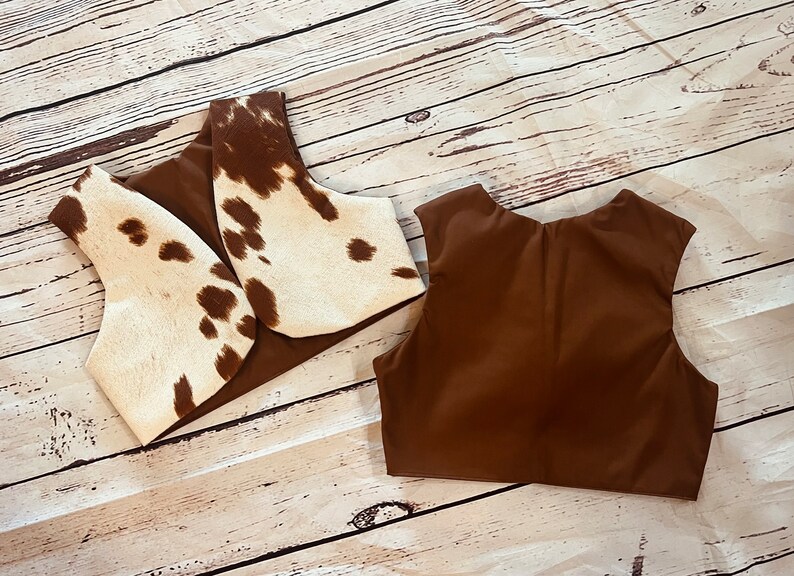 May include: Two brown vests, one with a white and brown cowhide pattern. The vests are laid flat on a light brown wooden surface.