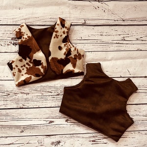 May include: Two brown and white cow print fabric vests. One vest is brown suede and the other is brown suede with a cow print fabric overlay.