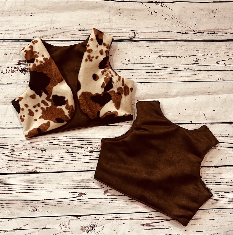 May include: Two brown and white cow print fabric vests. One vest is brown suede and the other is brown suede with a cow print fabric overlay.