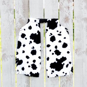 May include: A black and white cow print chaps with fringe. The chaps are hanging on a white wooden fence.
